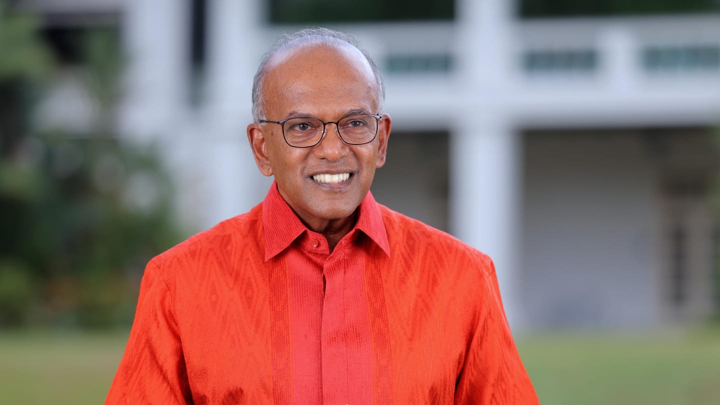 Close-up of man in orange patterned shirt; white building behind.